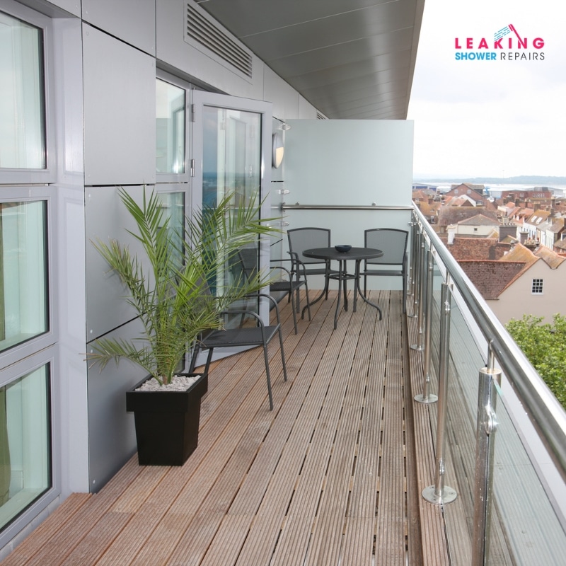 Modern balcony with glass railing, wooden deck, and outdoor seating showing signs of water damage.