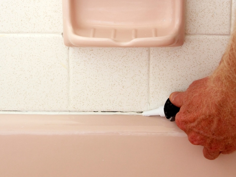 Person sealing gaps around a bathtub to prevent cracked grout in the tile joints from water damage.