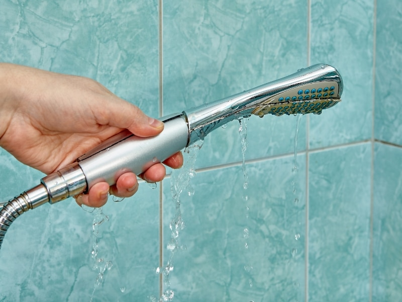 A hand holds a dripping shower head against tiled wall, suggesting diy shower leak repairs.