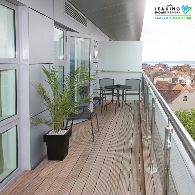 Modern balcony with glass railing, wooden deck, and outdoor seating showing signs of water damage.